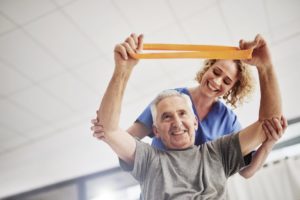 A sminling senior women lifting dumbbells above her head with the assistance of a smiling therapist who has her hands on the woman's elbows to assist with the lift.